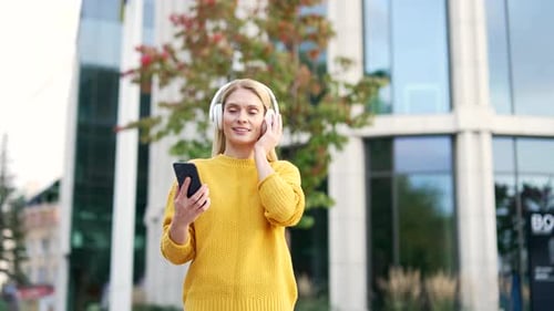 Happy smiling woman listens to music on wireless headphones while walking on the street near