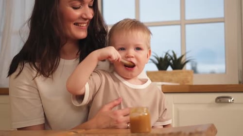 Mother Watching Blond Toddler Eat Food in Kitchen