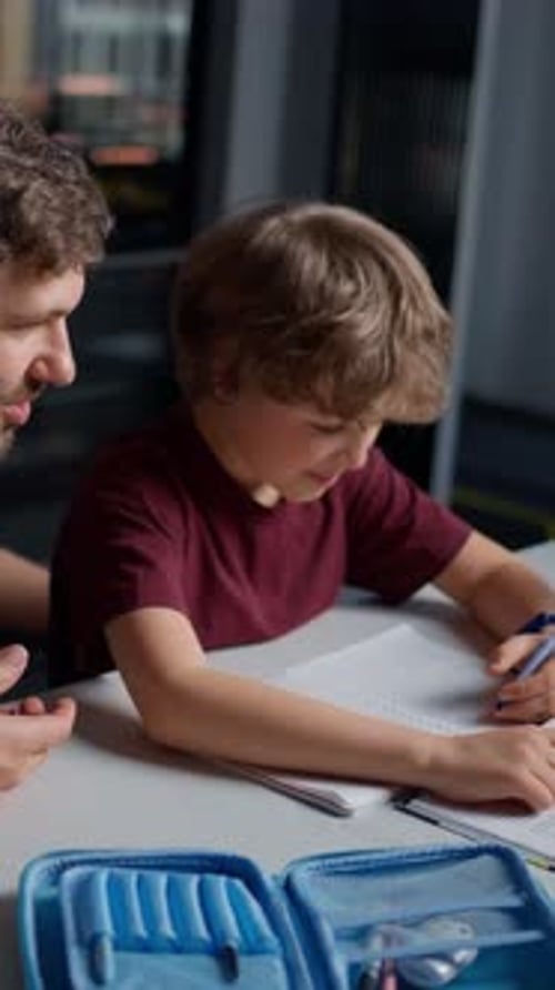 Young Boy Focused on Writing in a Notebook While Receiving Guidance and Support From an Adult Male