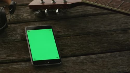Telefone com tela verde no modo retrato colocado em uma mesa de madeira ao lado de guitarra e fones de ouvido.