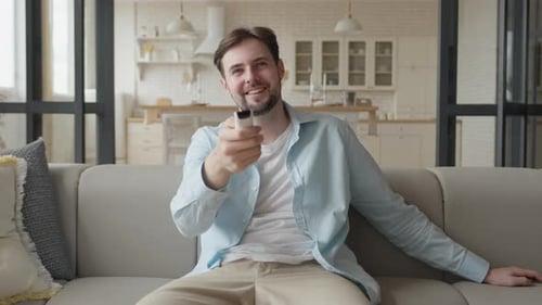 Smiling Man Points Remote on Couch at Camera
