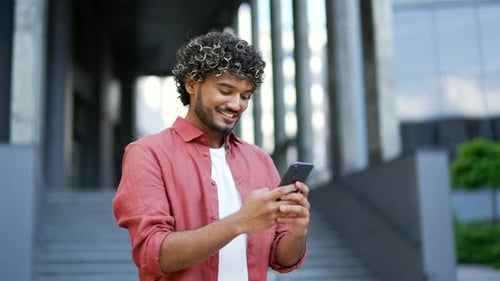 Young smiling businessman uses a mobile phone standing on street near office building. Handsome male