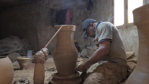 Potter giving final touches to the large clay vase on potters wheel. Side view.