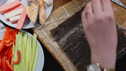 Top down view of hands placing Nori on top of wooden board, adding rice