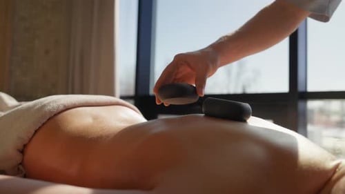 Entspannende Hot-Stone-Therapie-Sitzung mit glatten schwarzen Steinen, die entlang einer Frau mit nacktem Rücken in einem