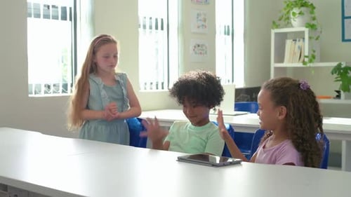 Diverse kids at school table, girl in blue dress approaching, initiating playful game, using tablet