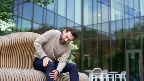 Businessman suffers from painful severe knee joint pain sitting on a bench on the street near