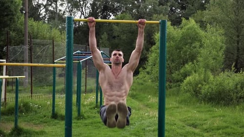 Athletic Man Trains Abdominal Muscles Raising Legs on Horizontal Bar at Sports Ground Young