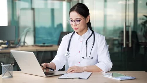 Young asian female doctor in a white coat writes a prescription using a laptop while sitting