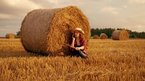 Woman Farm Worker in Rubber Boots Seating at Round Hay Bale in Harvested Agricultural Field Female