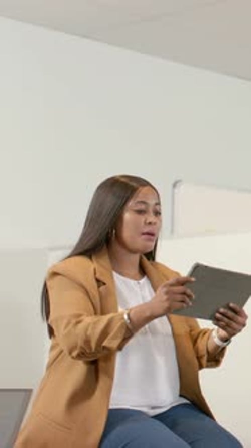 Vertical video: Raising midadult woman in tan blazer tapping tablet while teaching at office laptop