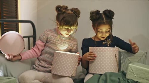 Two Young Girls Sitting on Bed and Opening Boxes with Presents Glowing From Inside Enjoying Their
