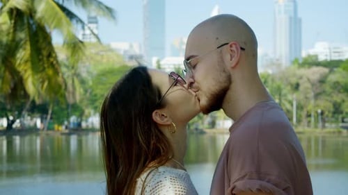 Wide Side View Captures Man and Woman Sharing a Kiss Outdoors Park Greenery and Distant Highrise