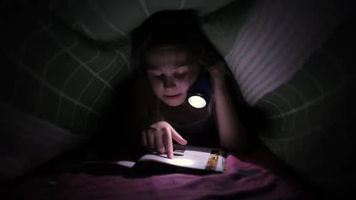 Little Child Girl Reading a Book with a Flashlight Under the Cover in Bed