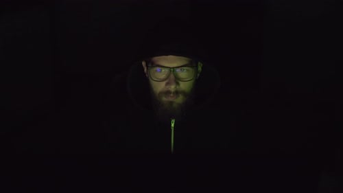 Ein Hacker mit unbedecktem Gesicht arbeitet am PC im dunklen Raum.