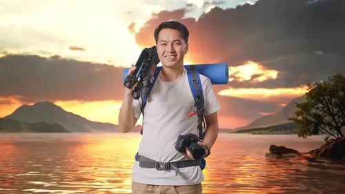 Asian Male Hiker Smiling, Holding A Camera, And Putting A Tripod On His Shoulder At A Lake