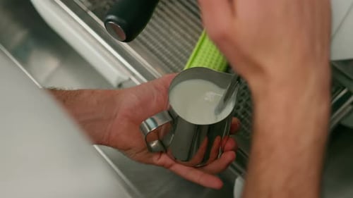barista uses coffee machine and churns fresh milk to prepare latte or cappuccino in cafe for client