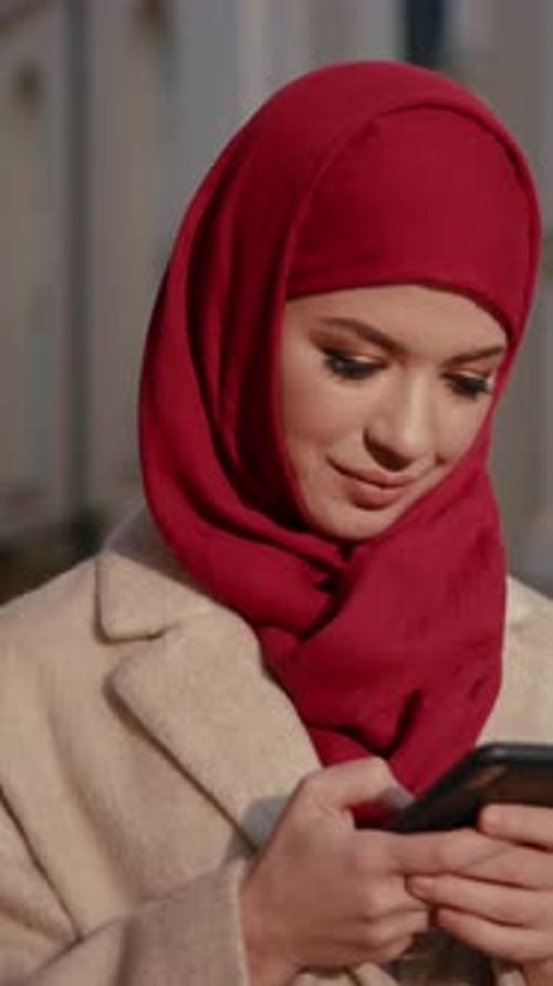 A Young Woman Dressed in a Vibrant Red Hijab is Actively Using Her Smartphone Outdoors