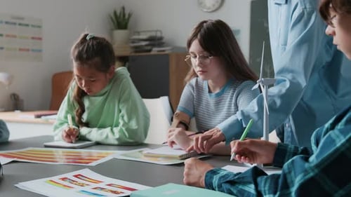 Children Working on Drawings Alongside Wind Turbine Model, Guided by Teacher