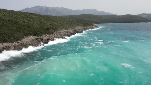 A Drone Flies Over a Rocky Coastline with a Bright Blue Sea Wave and Mountains in the Background