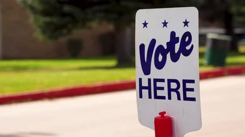 Blue Vote Here Sign Close up with People Driving Cars Walking in the Background