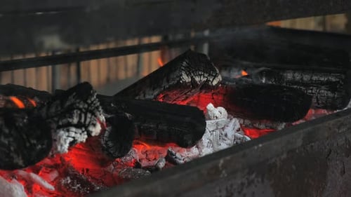 Pfanne in Zeitlupe entlang eines Metallgrills mit offener orangefarbener, heißer Feuerflamme, die flackert, während Asche und Glut rinnen
