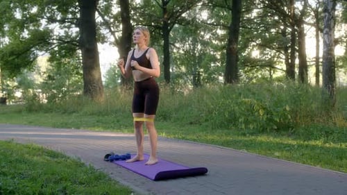 Slender Athletic Girl in Black Tightfitting Tracksuit Does Squats with Elastic Band on Street in