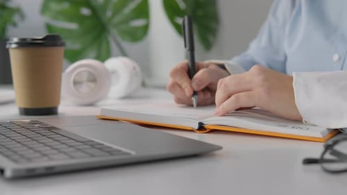 Person Writing in Notebook at Desk with Laptop