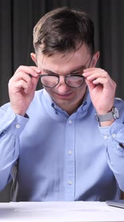 Vertical Video Tired Office Worker Rubbing Eyes at Desk with Folders and Laptop