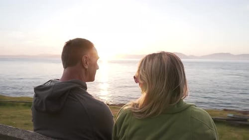 Beautiful adult family kiss while watching sunset on coastline bench, motion view