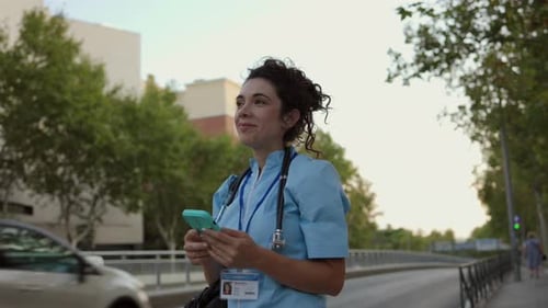 A young nurse in scrubs checks her smartphone while walking outdoors, smiling and relaxed after work