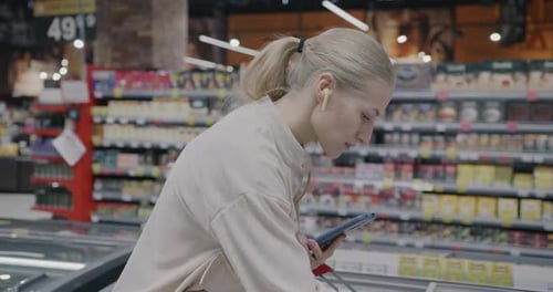 Young Woman Wearing Earphones Using Smartphone and Checking Groceries in Trolley in Supermarket