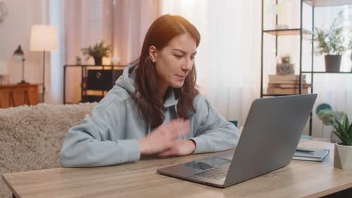 Glückliche junge Frau sitzt am Tisch und spricht per Videoanruf per Laptop zu Hause mit der Hand