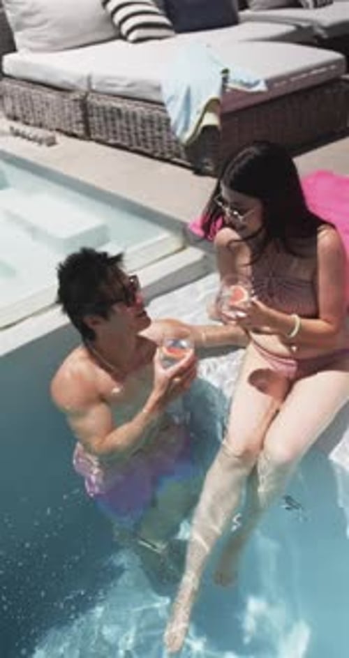 Vertical video: Raising man in trunks offering drinks to woman in pink swimwear at pool, toasting