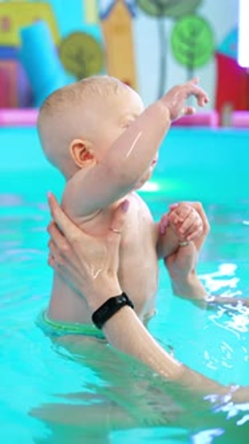 Female hands hold the adorable blond baby in the swimming pool. Lovely child looks up with interest.