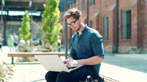 Trendy handsome caucasian man using laptop outdoors, working online in urban city area on a bench.