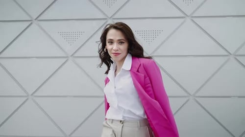 A Cheerful Girl in a Pink Jacket and Light Trousers Walks Around the Business Center with a White