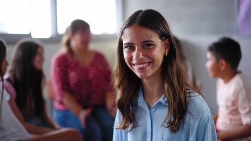 Primary School Girl Poses Looking Smiling at Camera Sitting in Class with Classmates in Background