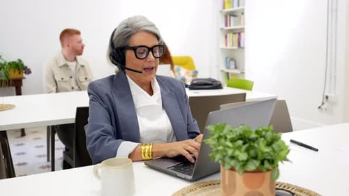 Senior Woman Talks with Headset Typing on Laptop