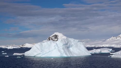 Im Meer befindet sich eine hellblaue Eisscholle. Auf dem Eisberg im Hintergrund sind einige Felsen freigelegt
.