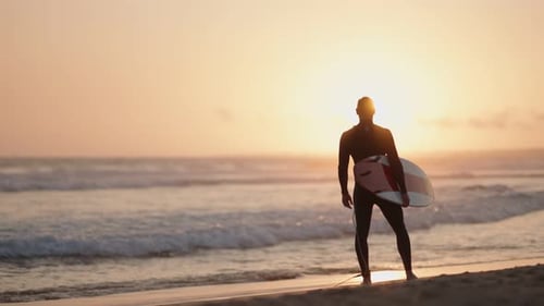 Surfer hält Surfbrett am Strand und beobachtet Meereswellen bei Sonnenuntergang