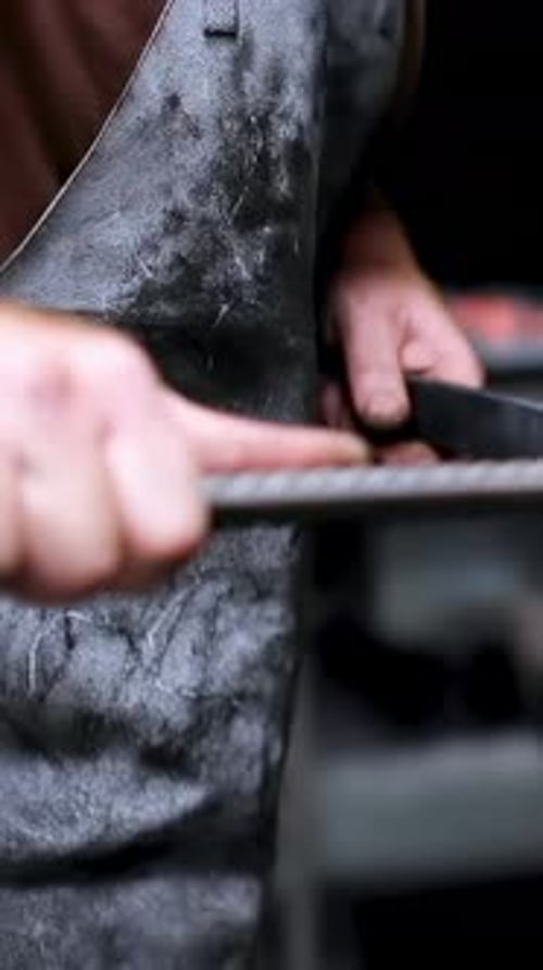 Vertical video: Gripping metal bar, man filing with rasp to shape metal at shop, in leather apron