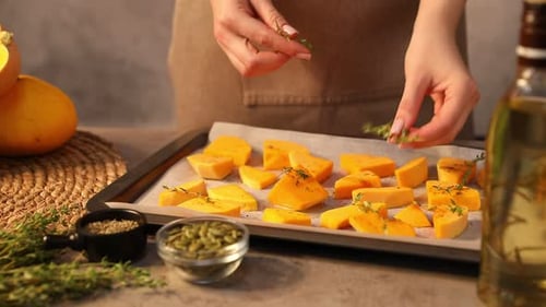 Woman adding thyme to pumpkin in baking tray at table, closeup