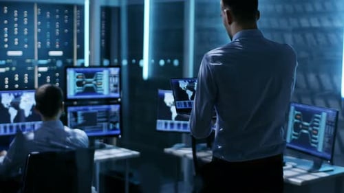 Im Systemüberwachungsraum nähert sich ein Techniker mit Laptop einem Kollegen, der an seinem Arbeitsplatz ist