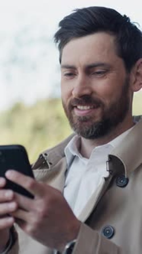 Smiling Young Stylish Handsome Man with Beard in Coat Uses Smartphone Stand in Part at Sunlight