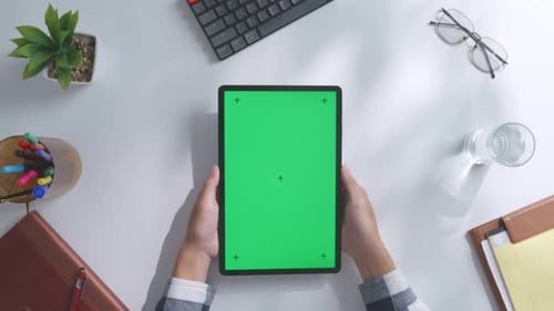Top down view of hands holding a mock up green screen tablet on a white desk