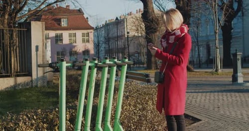 Young Blonde Woman Using Smartphone While Unlocking Green Escooter on Street
