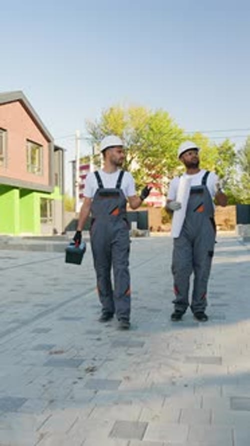 Dos constructores con cascos y ropa de trabajo caminan cerca de una moderna casa adosada verde y de ladrillo hablando sobre