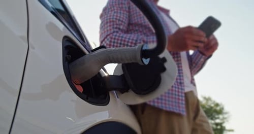 Man With Mobile Phone Stands Near Electric Car Connected to a Charging Station