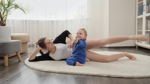 Cute baby boy watching young mother lying on floor and doing stretching and practising fitness